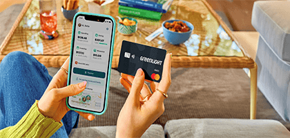 A person sitting indoors holds a smartphone in one hand displaying the Greenlight app with spending, saving, and investing balances, and a black Greenlight debit card in the other. A coffee table with snacks, books, and a drink is visible in the background, creating a casual home setting.