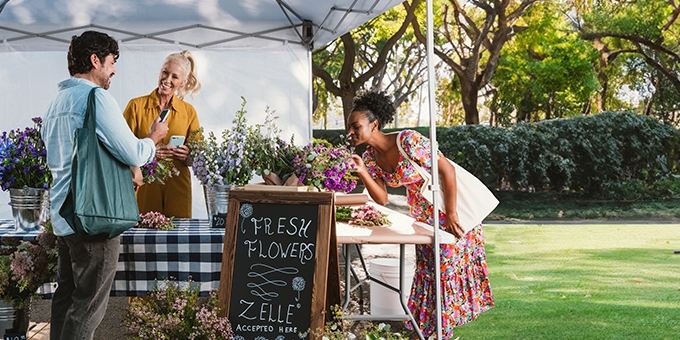 People buying flowers at an outdoor booth with a Zelle accepted here sign.