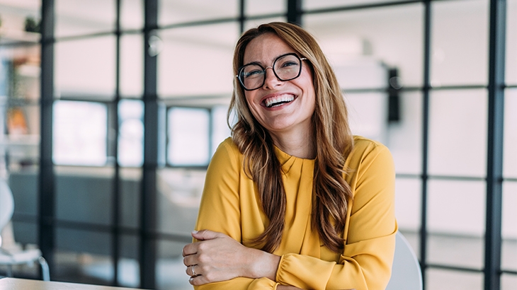 A blonde woman with glasses in a yellow top is sitting at a desk and smiling broadly.
