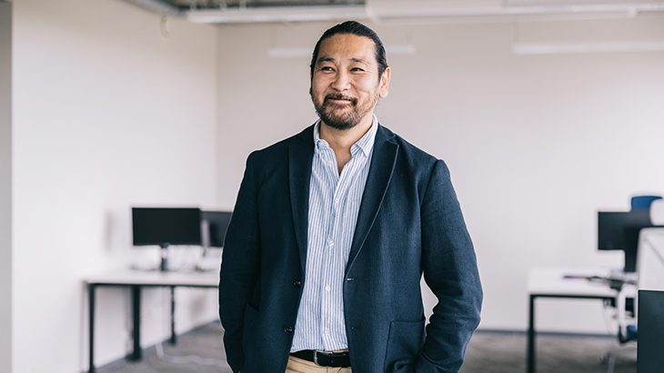 A smiling professional man in a navy blazer stands in a modern, open office.