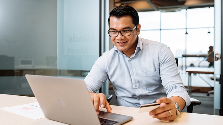A smiling man with glasses, wearing a light blue shirt, sits at a desk using a laptop and holding a debit or credit card.