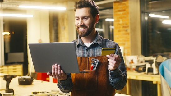 A smiling male craftsman with a beard, wearing a plaid shirt and a leather workshop apron, holds a laptop in one hand and a gold credit card in the other.