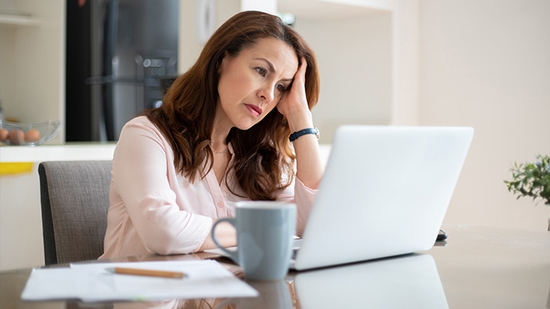 A stressed and worried woman resting her head in her hand while looking closely at her laptop screen.