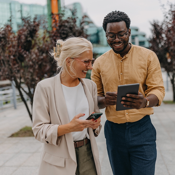 Two smiling professionals standing outdoors, looking at a tablet together.