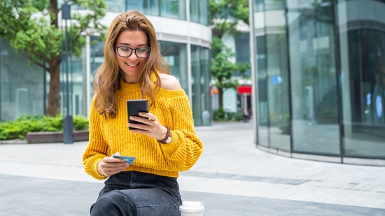 Mujer sonriente usa la app móvil de WaFd Bank en su smartphone y sostiene una tarjeta de crédito al aire libre.