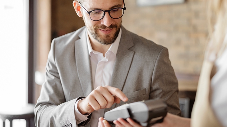 A smiling man with a beard and glasses, wearing a light gray suit jacket, is tapping on a payment terminal held by an unseen person.