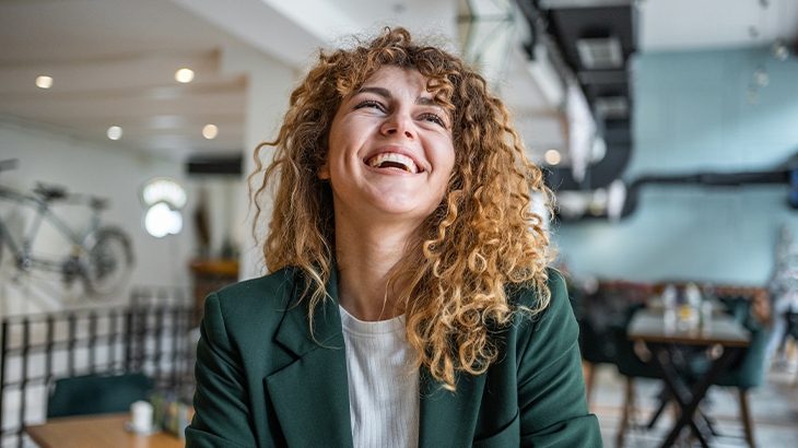 A happy, curly-haired woman in a green blazer is smiling and looking up while sitting in a cafe.