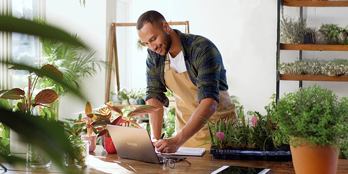 A business owner in an apron working on a laptop at a wooden table in a plant-filled shop or nursery.