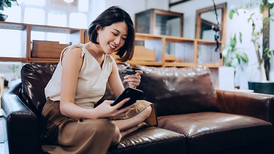 Una mujer sonriendo sentada en un sofá viendo su tarjeta de crédito y su teléfono.