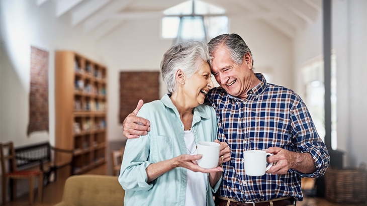 Mature couple in their living room hugging and holding mugs.