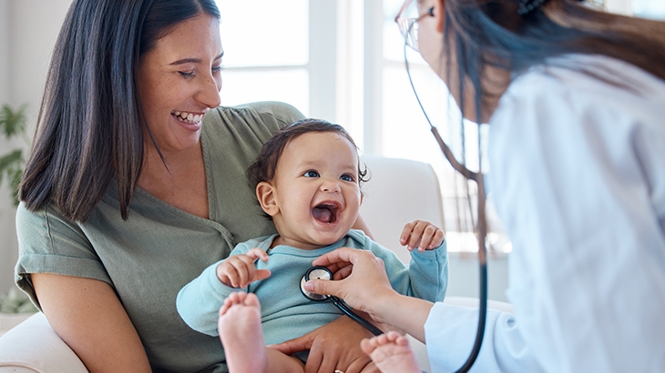 Baby on mother's lap smiling and getting examined by doctor.