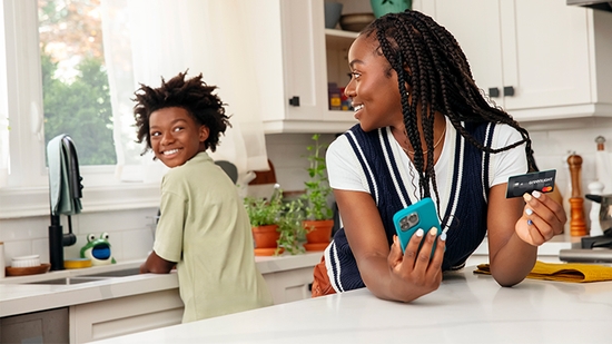 Siblings in the kitchen, one holding mobile phone and a Greenlight debit card.