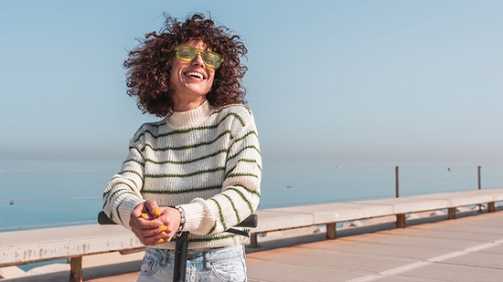 A woman with curly hair and yellow sunglasses smiles while leaning on the handlebars of a scooter near the ocean.