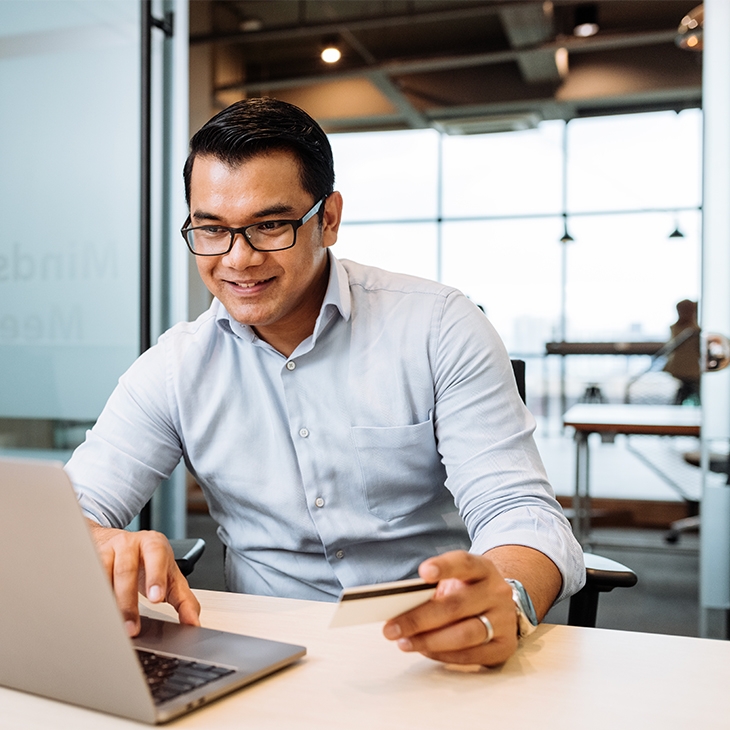 A smiling man with glasses, wearing a light blue shirt, sits at a desk using a laptop and holding a debit or credit card.