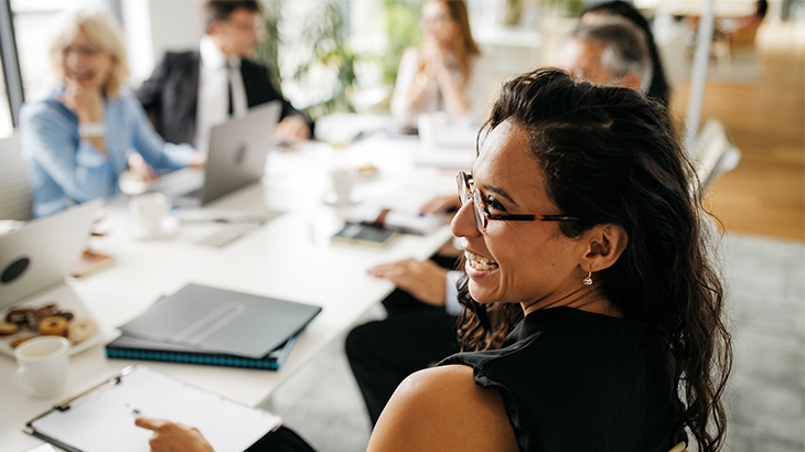 A professional woman with dark hair and glasses, wearing a black top, smiles while sitting at a conference table with a group of colleagues in a bright office.