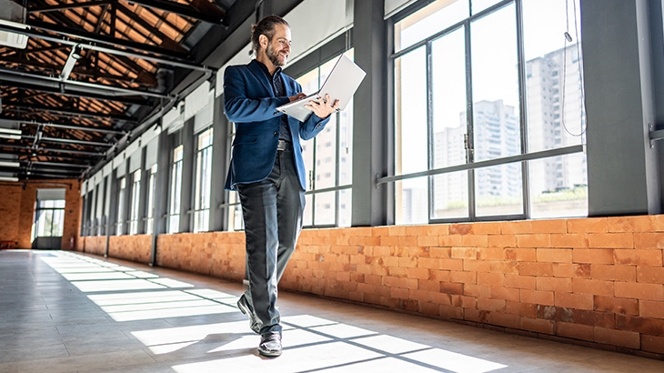 Man walking while holding laptop through empty commercial building hallway.