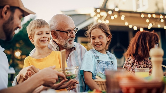 Una familia multigeneracional sonríe y conversa mientras comparte una comida en un patio al aire libre decorado con luces.