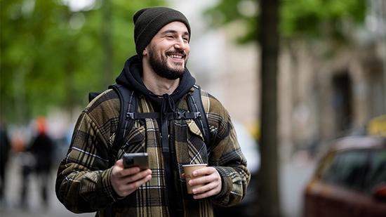 Un hombre sonriente con barba, que lleva una chaqueta de cuadros y un gorro, camina por la calle sosteniendo un smartphone y una taza de café.