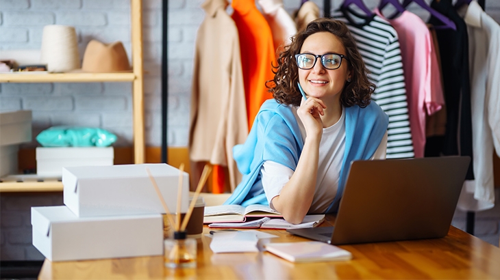 A smiling woman working on her laptop in a clothing shop.