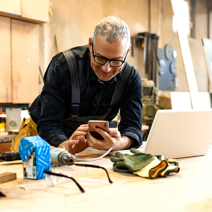 A man in a workshop wearing glasses and overalls smiles while looking at his smartphone, leaning over a workbench with a laptop, tools, and safety gear.