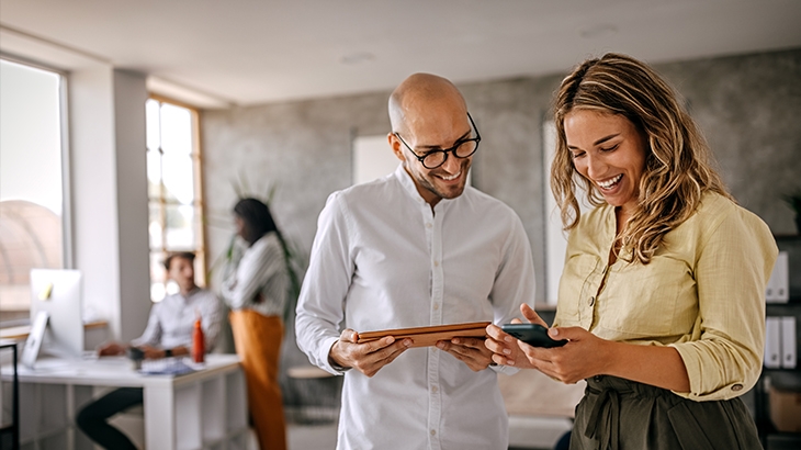 Two smiling professionals, a man and a woman, look at a tablet and a phone while standing in a busy, modern office.