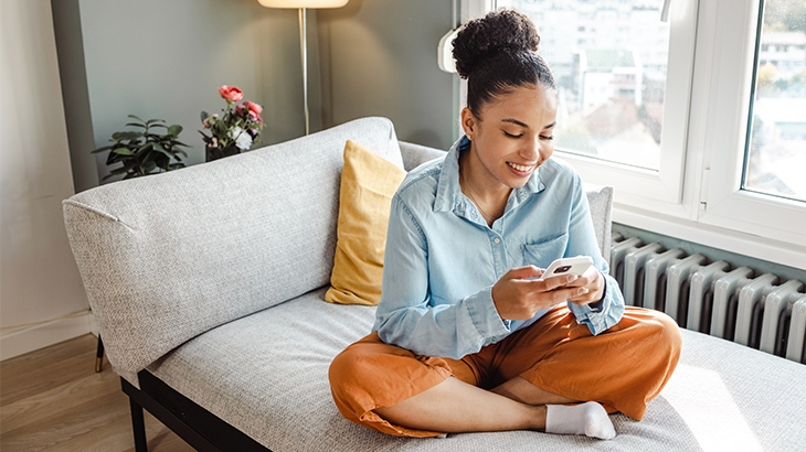 Mujer usa su celular mientras está sentada con las piernas cruzadas en un sofá.