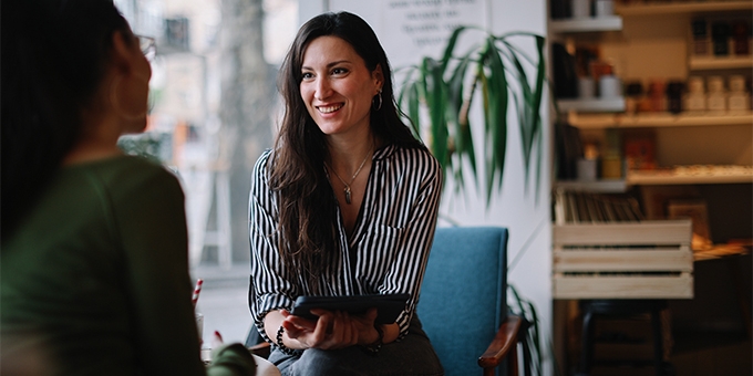 A woman in a striped shirt smiles while holding a tablet and talking to a colleague in a bright, modern cafe setting.