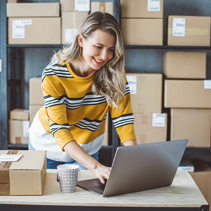 Woman in a yellow and black striped sweater works on a laptop surrounded by cardboard shipping boxes in a small business or warehouse.