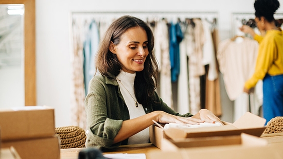A smiling small business owner carefully packing an order into a cardboard shipping box inside a clothing boutique.