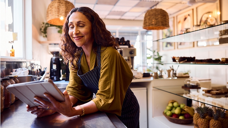 A smiling female small business owner wearing an apron is leaning on a counter and looking down at a digital tablet in her hands.