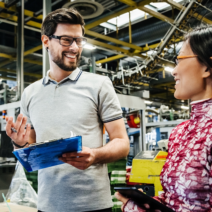 Two business colleagues smiling and talking in a factory or warehouse setting, with the man holding a clipboard.