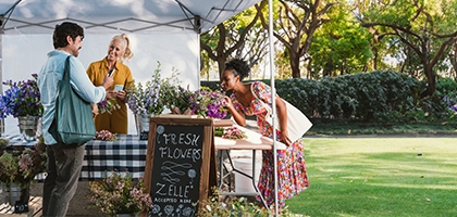 People buying flowers at an outdoor booth with a Zelle accepted here sign.