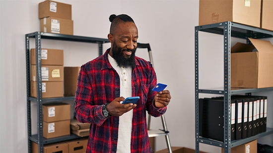 A man wearing a red plaid shirt smiles while holding a blue credit card and a smartphone in a warehouse filled with shipping boxes.