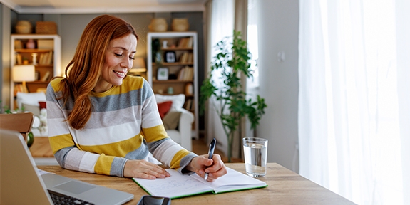 Woman seated at a dining table, planning her time with a notebook and laptop.