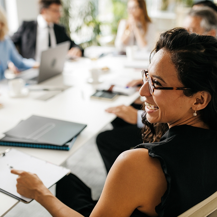 A professional woman with dark hair and glasses, wearing a black top, smiles while sitting at a conference table with a group of colleagues in a bright office.