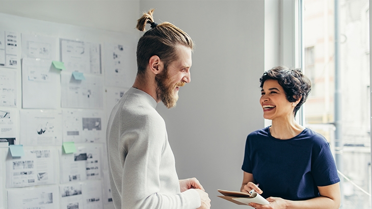 Two smiling colleagues discussing work with documents on a wall in the background.