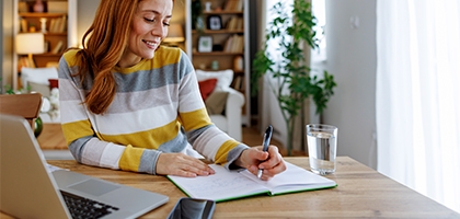 Woman seated at a dining table, planning her time with a notebook and laptop.