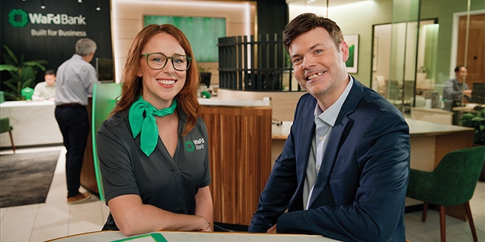 A smiling woman in a WaFd Bank uniform and a man in a blue suit sitting together inside a modern WaFd Bank branch.