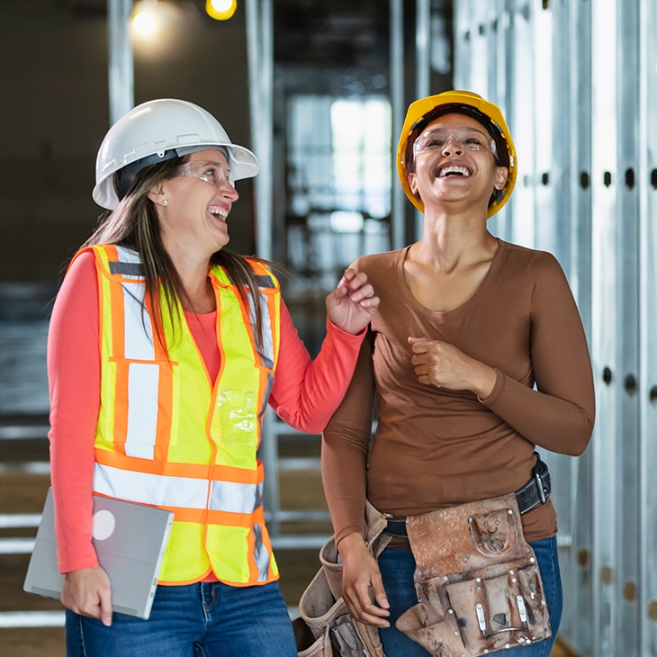 Two women in hard hats and safety gear laugh together while walking through a building under construction with metal wall framing.