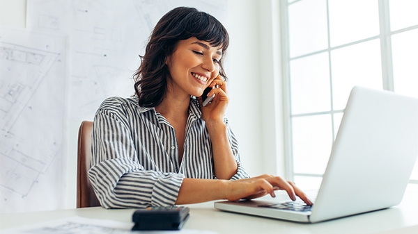 Businesswoman in office smiling while on the phone and using a laptop.