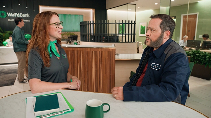 A WaFd Bank employee with red hair smiles and talks to a customer in a work jacket at a modern branch counter.