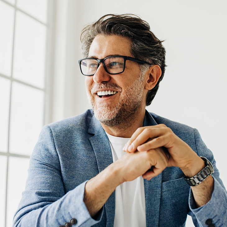 A smiling, middle-aged man with graying hair and a beard, wearing glasses and a light blue blazer, sits in front of a large window.