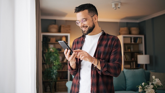 A man with a beard and glasses looks at his smartphone and smiles while standing near a window.
