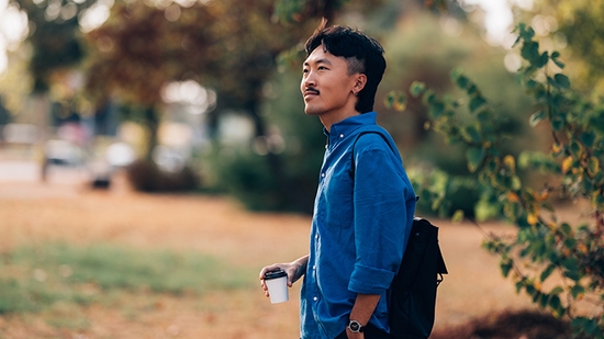 A man in a blue shirt holding a coffee cup while standing outdoors in a park setting.