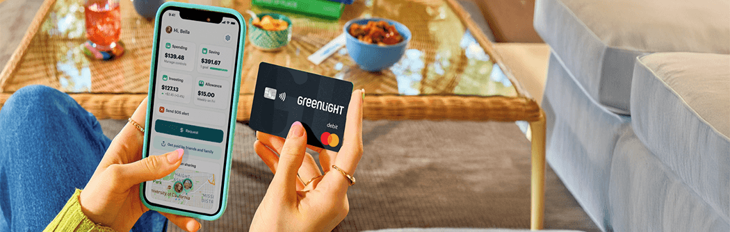 A person sitting indoors holds a smartphone in one hand displaying the Greenlight app with spending, saving, and investing balances, and a black Greenlight debit card in the other. A coffee table with snacks, books, and a drink is visible in the background, creating a casual home setting.