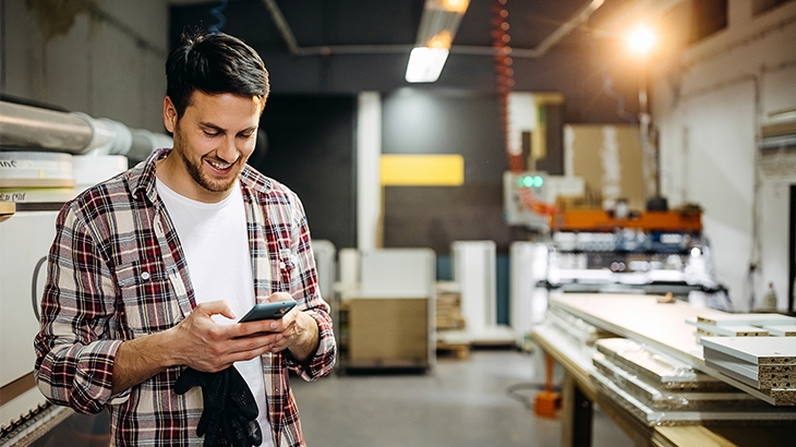 A smiling man in a plaid shirt and white t-shirt stands in a workshop, looking down at his phone.