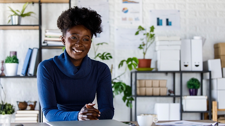 A young business woman in a turtleneck sitting in her office smiling.