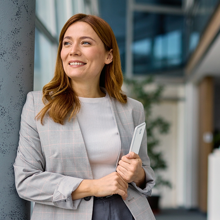 A confident, smiling woman in a light gray blazer, holding a tablet, stands in a modern, brightly lit office looking toward the window.