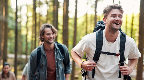 A group of friends with backpacks smiling and laughing while hiking together through a sunlit forest trail.