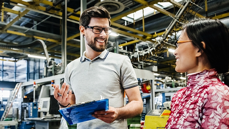 Two business colleagues smiling and talking in a factory or warehouse setting, with the man holding a clipboard.